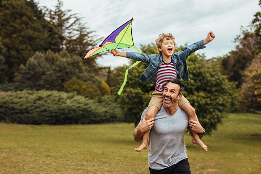 Father carrying son playing with kite in park. Excited little boy sits on shoulders of dad holding a kite.