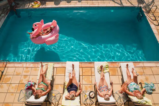 Group of senior women relaxing by the poolside while on holiday. There are four women laid on sun beds and then one woman laid on an inflatable flamingo on the pool water.