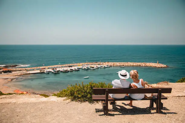 Senior couple sitting down on a bench overlooking the Mediterranean Sea. They are relaxing and spending time together.
