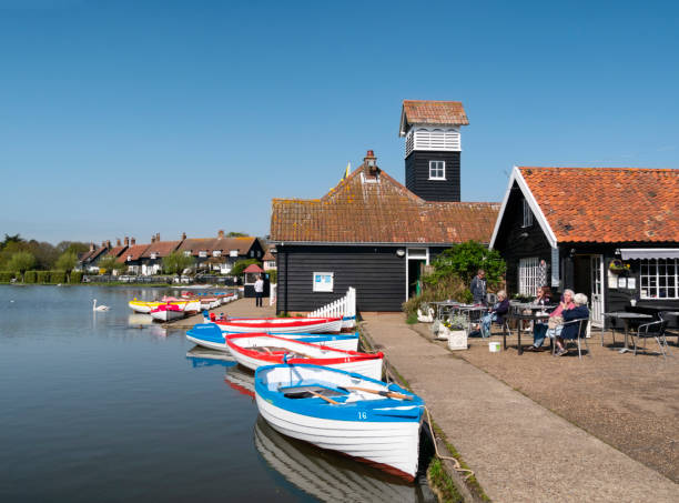 People sitting outside a tearoom near a cluster of rowing boats at Thorpeness Meare in Suffolk, Eastern England. Thorpeness was developed into a ‘fantasy’ village by the landowner, Glencairn Stuart Ogilvie, and included a man-made mere - known as The Meare - and a set of almshouses as well as many picturesque houses. One of the village’s most notable features is a water tower (not seen here) which was disguised as a very tall house: the water tower is now a residential building. Every August, Thorpeness Meare is the setting for the Thorpeness Regatta.