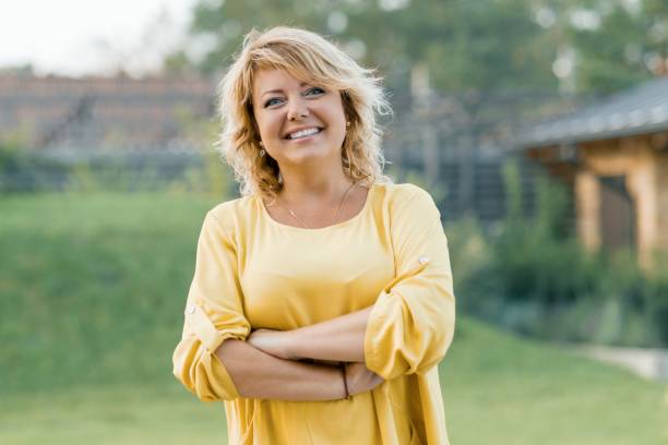 portrait en plein air de femme mûre confiante positive. souriante femme blonde vêtue d’une robe jaune avec les bras croisés près de la maison - 45 49 ans photos et images de collection