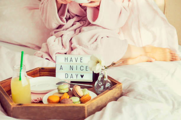 istock Good morning concept. Breakfast in bed with Have a nice day text on lighted box, juice and macaroons on tray and blurred woman in bathrobe drinking coffee. Hospitality, care, service. Copy space. 1078667682