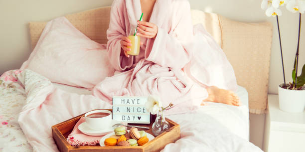 istock Good morning concept. Breakfast in bed with Have a nice day text on lighted box, coffee and macaroons on tray and blurred woman in bathrobe drinking juice. Hospitality, care, service. Copy space. 1078691820