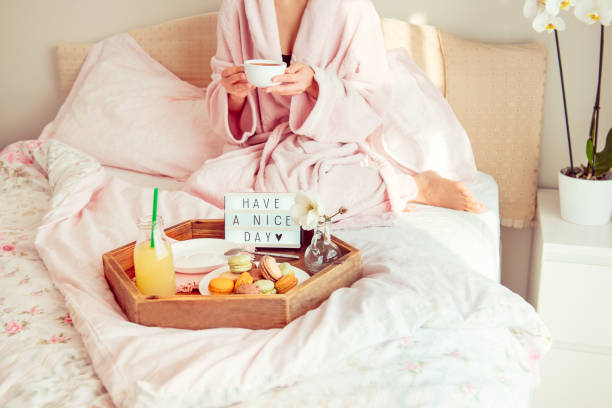 istock Good morning concept. Breakfast in bed with Have a nice day text on lighted box, juice and macaroons on tray and blurred woman in bathrobe drinking coffee. Hospitality, care, service. Copy space. 1078691822