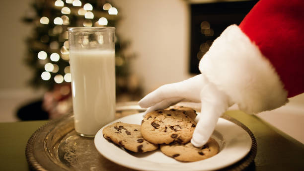 la mano guantata di babbo natale raccoglie un biscotto di cioccolato da un vassoio con un bicchiere di latte su di esso con un albero di natale e un camino sullo sfondo la vigilia di natale - gloved foto e immagini stock