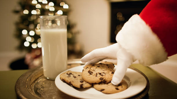 サンタクロースの手袋の手は、クリスマスイブに背景にクリスマスツリーと暖炉とミルクのグラスでトレイからチョコレートチップクッキーを拾います - gloved ストックフォトと画像