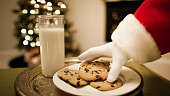 Santa Claus' Gloved Hand Picks Up a Chocolate Chip Cookie from a Tray with a Glass of Milk on It with a Christmas Tree and a Fireplace in the Background on Christmas Eve