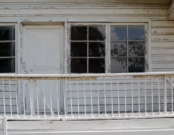 A white, abandoned building with three glass pane windows, a door, and a bowed railing.
