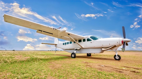 A small white-colored propeller airplane sits alone on a dirt and grass landing strip in wide open African bush land, with a vibrant blue sky in the background.