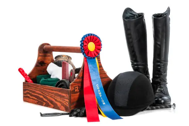 A multi-colored equine Champion award ribbon clipped on a wooden groom's tote with a pair of shiny black leather tall riding boots, helmet, rising crop and gloves sitting on a white background.