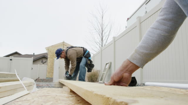 A Latino Man in His Forties Uses a Circular Saw to Cut a Wooden Plank as Another Person Secures the Board on a Construction Site in Winter Under an Overcast Sky