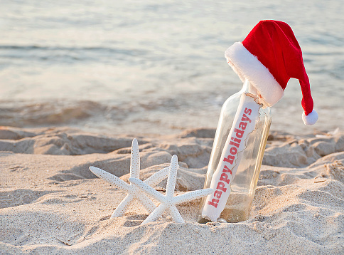 Santa hat on message in a bottle with pair of white starfish in beach sand and Happy Holidays greeting