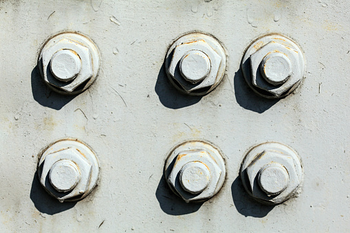 Six large nuts and bolts on gray steel plate of rail bridge, lit by bright sun. Abstract industrial background.