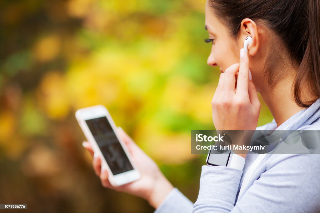 Photo of joyful fitness woman 20s in sportswear touching bluetooth earpod and holding mobile phone, while resting in green park. Headphones Stock Photo