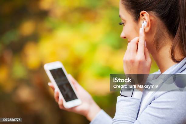 Photo Of Joyful Fitness Woman 20s In Sportswear Touching Bluetooth Earpod And Holding Mobile Phone While Resting In Green Park Stock Photo - Download Image Now