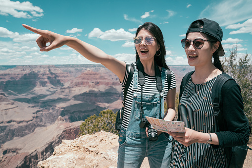 happy women travelers pointing to the destination standing on the skyline in grand canyon national park. young sisters travel with paper map on summer holidays. tour in america lifestyle concept.