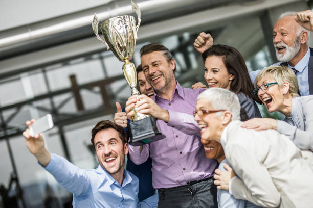 Selfie with a trophy! Large group of happy business people having fun while taking a selfie with winning trophy in the office. office competition stock pictures, royalty-free photos & images