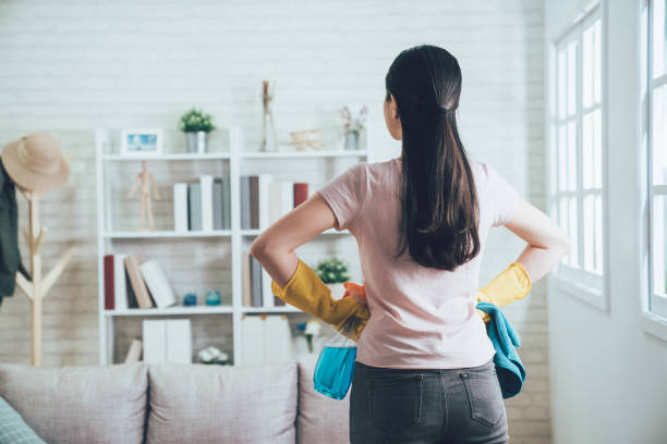 housekeeper looking at the clean living room back view of asian housekeeper looking at the clean living room after she tidied up. young wife finished house chores putting hands in waist watching the bookshelf beside the sunlight window. housework stock pictures, royalty-free photos & images