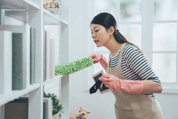 asian housekeeper in apron dusting the bookshelf asian housekeeper in apron dusting the bookshelf by feather duster taking up the picture frame carefully cleaning in living room at home. young wife in rubber gloves doing housework. dusting stock pictures, royalty-free photos & images