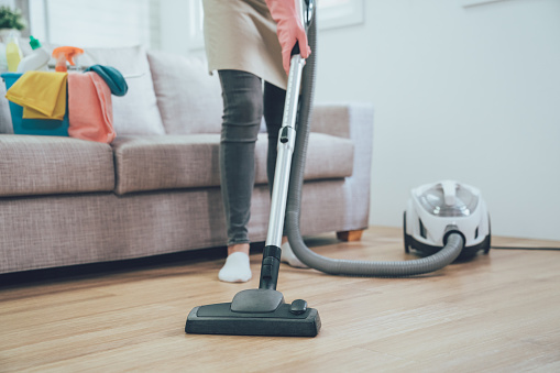 asian woman using vacuum cleaner in the living room. housekeeper wearing pink protective rubber gloves cleaning the wooden floor in the house. full of clean supplies in bucket on comfortable sofa