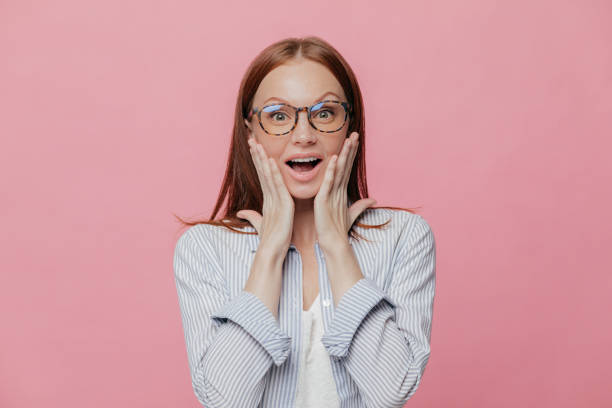 istock Omg, how nice! Beautiful elegant female employee touches cheeks with both palms, wears spectacles, elegant shirt, cant believe in her promotion, recieves reward, isolated over pink background 1079594496