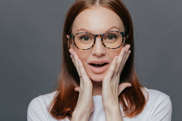 istock Headshot of surprised young Caucasian lady keeps both palms on her cheeks, looks with amazement, recieves unexpected news, wears spectacles, white clothing, poses alone against grey background 1079594746