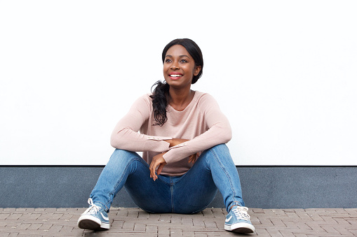 Portrait of young black woman sitting on floor by white wall and smiling