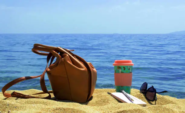 Photo of Backpack, tumbler cup, sunglasses and notebook on the sand by the sea. Tourist objects on holiday.