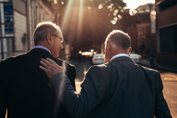 Senior businessman walking together outdoors Rear view of two senior businessman walking together outdoors on city street. Mature businessman with hand in back of male colleague walking outdoors on a sunny day. side by side stock pictures, royalty-free photos & images