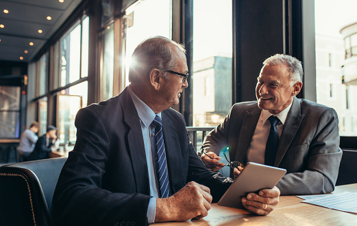 Two senior businessmen having an informal meeting at cafe. Businessman holding digital tablet talking with his male partner while sitting at modern coffee shop table.