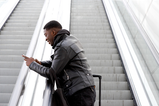 Portrait of cool young black man traveling with mobile phone and bag on escalator