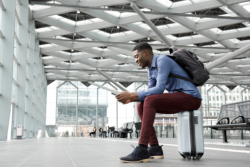Side portrait of young black man sitting on suitcase at station with mobile phone