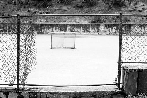 Black and white image of an opening in the wire fence is leading into an empty playground with a small mini soccer goal. The opening in the fence makes the opposite goal.