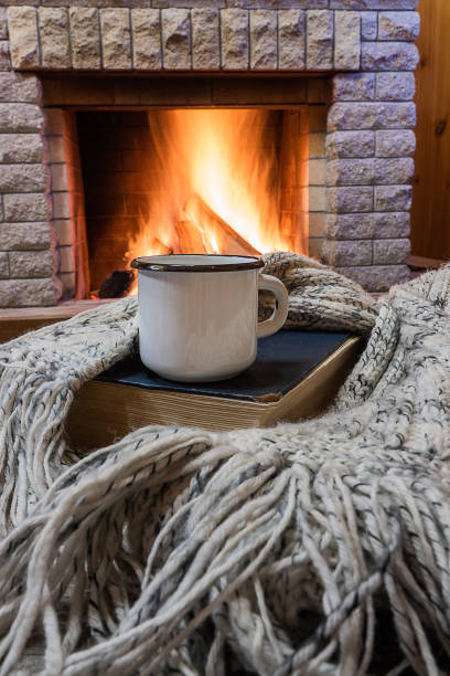 istock Cozy scene before fireplace with White enameled mug with tea, a book, and wool scarf. 1079756472