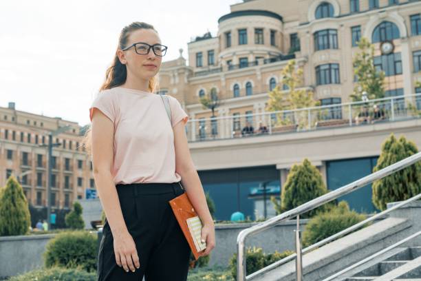 outdoor portrait of female student 16, 17 years old. girl in glasses, with backpack, textbooks. city background - 13 14 years teenager school education imagens e fotografias de stock