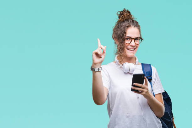 jovem estudante morena menina vestindo mochila, fones de ouvido e smartphone sobre fundo isolado surpreendido com um dedo apontando a ideia ou pergunta com cara feliz, número um - travel teenager talking student - fotografias e filmes do acervo