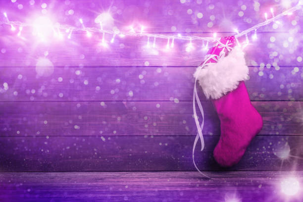 Pink Santa stocking with gift boxes hanging on glowing garland over holiday wooden background.