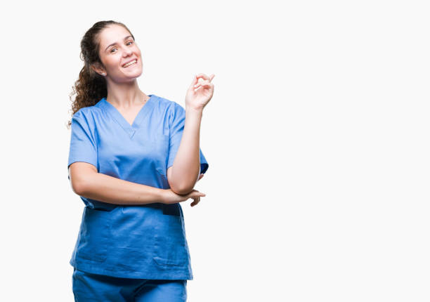 若いブルネットの医者女の子の顔に大きな笑みを浮かべて隔離された背景に看護師または外科医の制服を着てはカメラを見て側に手と指で指しています。 - welcome sign ストックフォトと画像
