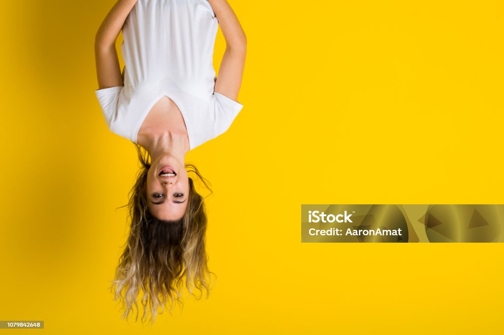 Beautiful young blonde woman jumping happy and excited hanging upside down over isolated yellow background Upside Down Stock Photo