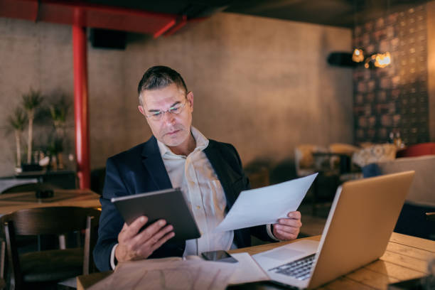 istock Caucasian businessman dressed smart casual holding tablet and paper and comparing data while sitting in cafe. On table laptop. 1079859248