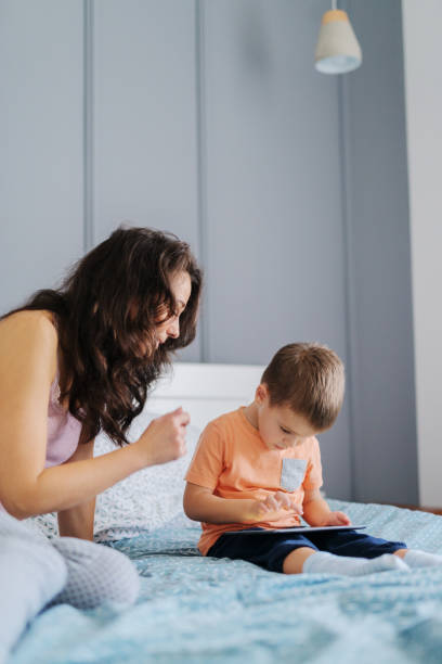 istock Little kid watching cartoons on tablet while sitting on the bed while his mother watching cartoons with him. Bedroom interior. 1079866428