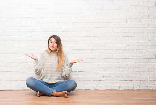 Young adult woman sitting on the floor over white brick wall at home clueless and confused expression with arms and hands raised. Doubt concept.