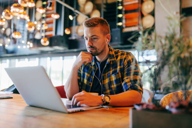 istock Young Caucasian bearded freelancer using laptop and sitting in cafe. One hand on keyboard and other on the chin. In ears earphones. Self employing concept. 1079891088