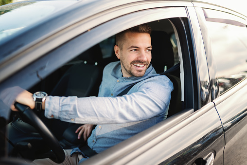 Smiling handsome Caucasian man driving his car and looking through window. Hand on steering wheel.