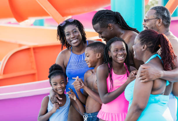 Multi-generation family at water park A multi-generation African-American family having fun together at a water park. The grandparents are in their 50s, the parents are in their 30s, and the children are 6 and 9 years old. They are standing in front of colorful water slides. The mother and 6 year old daughter are looking at the camera. male swimsuit standing arm around stock pictures, royalty-free photos & images