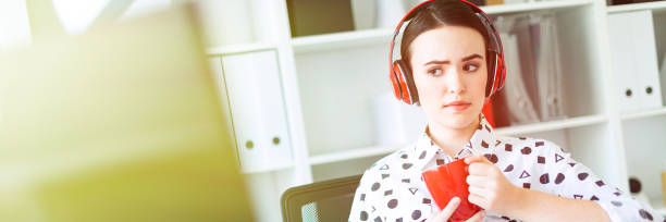 une jeune fille se trouve dans le casque à une table dans le bureau, tient une tasse rouge dans ses mains et se penche sur le moniteur. - the thinker audio photos et images de collection