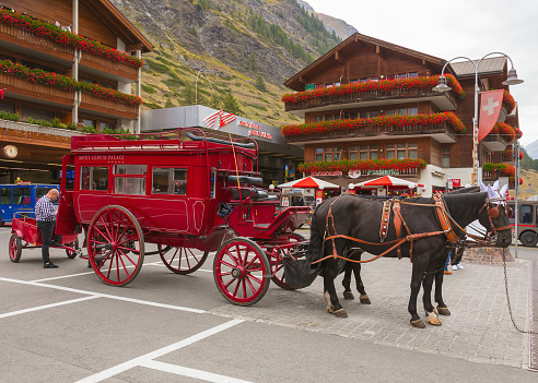 Zermatt, Switzerland - September 16, 2018: Bahnhofplatz square at the railway station in the town of Zermatt in the morning, a person standing at a horse-drawn carriage of the Hotel Mont Cervin Palace. Zermatt is a municipality in the district of Visp in the Swiss canton of Wallis, it is a famous mountaineering and ski resort.