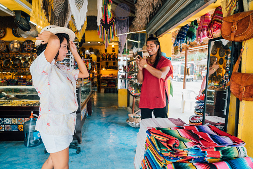 Adult Female trying on a Straw Hat while her friend takes a picture with Smart Phone in a touristic shop in Tulum