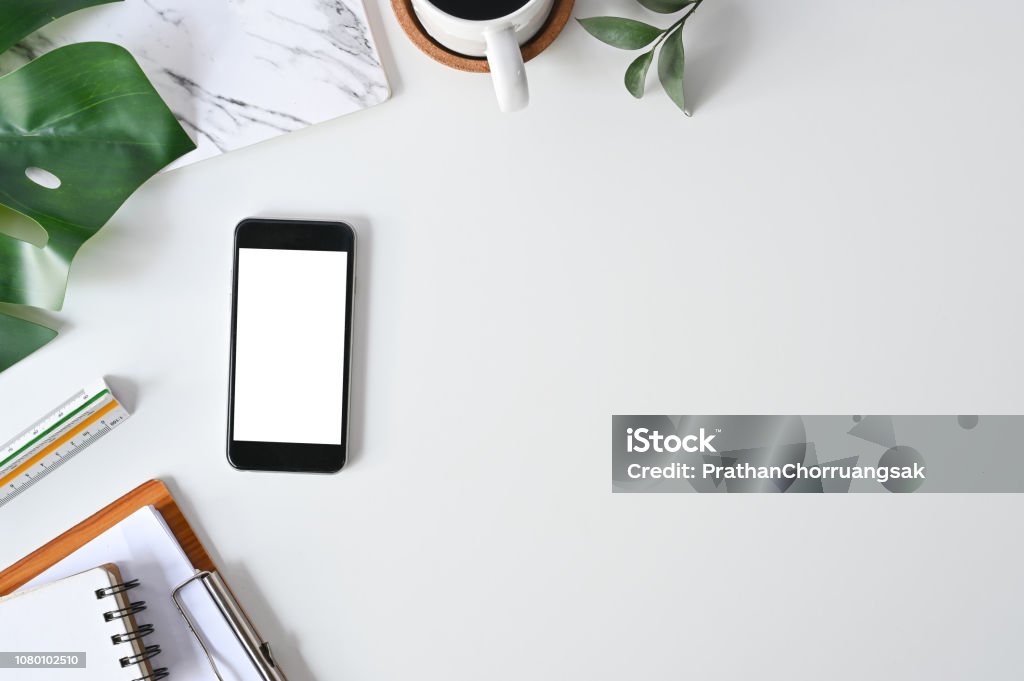 Top view office desk. Workspace with mockup smartphone, keyboard and office supplies, pencil, green leaf with coffee on white background. Telephone Stock Photo