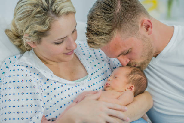 A man is sitting with his wife as she lies in a hospital bed. She is holding their newborn baby and he is kissing the baby's head.